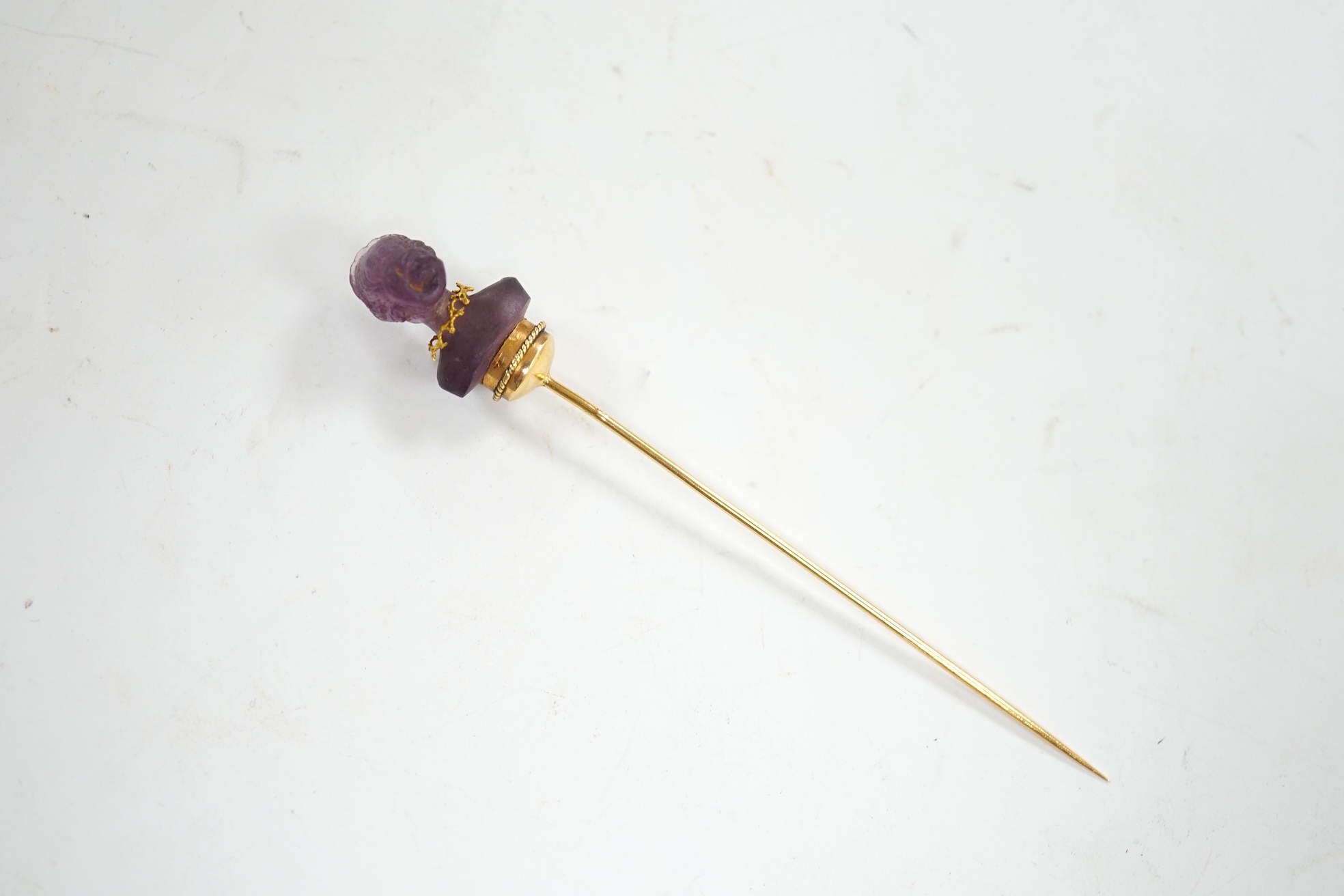 An early 20th century French yellow metal (18ct poincon mark) and amethyst paste set stick pin, the terminal modelled as the bust of a lady, 96mm, gross weight 5.6 grams. Condition - poor to fair